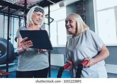 This Is So Funny. Smiling Senior Woman Asking Her Beaming Female Friend Working With Battle Ropes To Look At A Tablet Computer While Both Taking Exercise Class At A Gym.