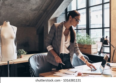 This is going to be a long day. Attractive young woman in eyewear using laptop while standing near the desk in her workshop 