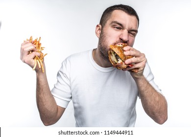 This Guy Is Very Delight Of Junk Food. He Is Biting A Big Piece Of Burger And Holding A Full Hand Of French Fries Covered With Ketchup. Young Amn Likes To Eat Oily Meal. Isolated On White Background.