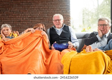 This heartwarming image captures a group of seniors sharing a moment of anticipation or suspense while watching TV together. They are cozily wrapped in a vivid orange blanket, with varied expressions - Powered by Shutterstock