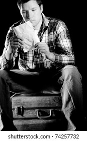 This Is A High Contrast, Black And White Image Of A Young Man Sitting On Some Old, Beat Up Suitcases Reading A Crumpled Up Letter. Shot With Hard Light On A Black Background.