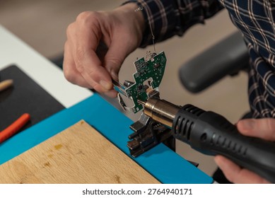 This high-angle close-up shot captures an electronics technician or engineer intently focused on repairing a small green printed circuit board. Using a precision hot air gun held in the right hand, the worker applies heat to specific components while stabilizing the PCB with the other hand. The workbench is equipped with various tools essential for micro-soldering and electronics repair, including orange-handled wire cutters, a cleaning brush, and a wooden soldering surface resting on a blue silicone mat. The blurred background reveals a modern workspace setup with computer monitors, suggesting a professional lab or home workshop dedicated to hardware development and repair.