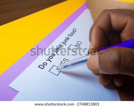 This high-angle, close-up shot captures a person’s hand poised to check a box on a survey. The hand, with a dark skin tone, holds a blue-capped ballpoint pen over a white paper containing the question, 