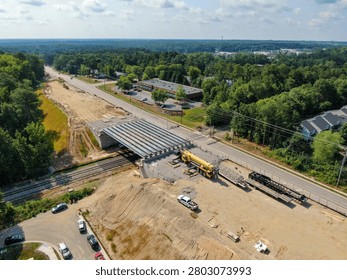 This high-angle drone photograph captures the dynamic infrastructure development at the S-Line passenger rail grade separation site in Raleigh, North Carolina. The image shows freshly positioned concrete bridge girders spanning across the roadway and adjacent railroad lines to eliminate an at-grade crossing. In the foreground, a large yellow mobile crane sits on a spacious dirt and gravel staging yard alongside supply trailers, framing a complete view of modern civil engineering and public transit expansion progress.