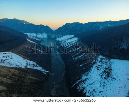 This high-angle drone shot captures the striking geography regarding a massive empty reservoir during the winter season. The deep valley features steep slopes with distinct horizontal terrace patterns formed by receding levels inside the golden-brown soil. A narrow silty river flows through the center, winding its way across the muddy basin. Thick forests featuring evergreen and deciduous trees line the upper ridges, contrasting with the barren landscape below. Pristine white snow patches are scattered across the valley floor and steep banks, adding bright highlights toward the earthy tones. In the background, rolling mountain ranges fade into a soft blue horizon under a clear cold sky. The scene emphasizes the immense scale and quiet isolation regarding this remote alpine wilderness. The intricate textures regarding eroded earth and the delicate stream create a rhythmic visual flow throughout the entire composition. This perspective highlights the dramatic environmental impact regarding seasonal changes inside the Romanian mountains.