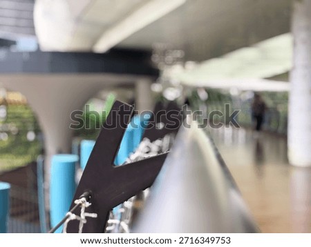 This high-angle, selective focus shot features a modern architectural railing or bridge structure. The foreground highlights a dark, angular metal bracket with thin cables, while the background transitions into a beautifully blurred (bokeh) urban walkway. The scene conveys a sense of contemporary design, transit, and city life with a clean, minimalist aesthetic.