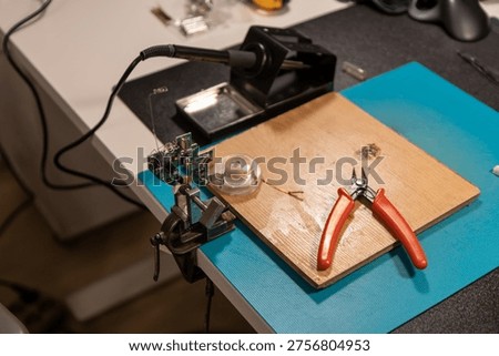 This high-angle shot captures a green printed circuit board (PCB) firmly gripped by the alligator clips of an adjustable third-hand soldering tool. The tool sits on a square piece of plywood, which shows signs of use with solder splashes. In the background, a soldering iron rests on a black work mat, and a teal silicone mat provides a non-slip surface for the repair work. The scene is illuminated by soft overhead lighting, highlighting the intricate components on the PCB and the metallic sheen of the helping hands. This image represents the meticulous process of electronics assembly, hobbyist tinkering, or professional circuit repair.