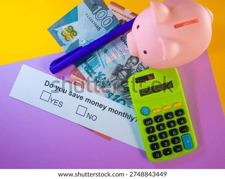 This high-angle shot features a pink piggy bank, a green calculator, and a blue pen arranged over Ghanaian Cedi banknotes. A white slip of paper with the text 