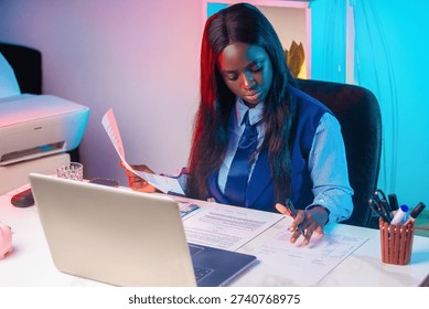 This high-angle shot features a young woman deeply engrossed in her work within a modern office setting. She is dressed in a professional blue collared shirt, tie, and vest. Her workspace is organized yet busy, featuring an open laptop, a white printer, and various documents spread across the desk. A striking dual-tone lighting scheme of neon pink and electric blue casts a creative, contemporary glow over the scene. She holds a pen and a sheet of paper, embodying productivity and concentration.