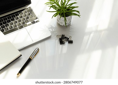 This high-angle, slightly diagonal shot captures a bright, modern workspace arranged on a glossy white desk. The top-left corner features a silver laptop, accompanied by a small green succulent in a geometric-patterned white pot and a small cluster of black binder clips. In the lower-left corner, a black and gold ballpoint pen lies next to the edge of an open notebook. Bright, natural sunlight streams across the scene from the right, casting sharp shadows and creating a clean, reflective backdrop. The lower-right area remains wide open, providing excellent copy space for business or productivity themes.