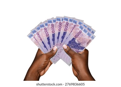 This high-angle studio shot features a pair of dark-skinned hands holding a fanned-out stack of West African CFA franc banknotes against a seamless white background. The currency shown is the 10,000 denomination ($XOF$), characterized by its vibrant purple and violet hues. The notes prominently display technical imagery, including a satellite dish and the iconic catfish-shaped gold weight. The composition emphasizes wealth, financial success, and regional African economic growth.
