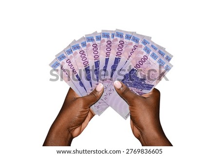 This high-angle studio shot features a pair of dark-skinned hands holding a fanned-out stack of West African CFA franc banknotes against a seamless white background. The currency shown is the 10,000 denomination ($XOF$), characterized by its vibrant purple and violet hues. The notes prominently display technical imagery, including a satellite dish and the iconic catfish-shaped gold weight. The composition emphasizes wealth, financial success, and regional African economic growth.