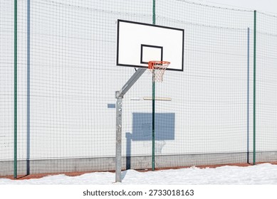 This high-angle, vertical shot captures a minimalist outdoor sports scene during the winter season. The central focus is a standard white rectangular basketball backboard with black markings and an orange metal rim with a white net. The hoop is mounted on a sturdy, weathered galvanized steel pole that shows signs of use and minor stickers. The entire structure is set against a backdrop of a clean, white safety mesh or netting, which creates a textured, grid-like pattern across the frame.

The ground is partially covered in crisp snow, contrasting with the dark reddish-brown pavement visible underneath. A distinct, sharp shadow of the basketball hoop is cast onto the white background by bright, direct sunlight, adding a layer of geometric depth to the composition. The lighting is cold and clear, emphasizing the lonely, quiet atmosphere of an urban sports ground during the off-season. The color palette is dominated by neutral whites, greys, and blacks, with the vibrant orange of the rim providing a singular pop of color.
