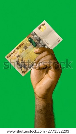 This high-angle, vertical shot features a Black person’s hand and forearm raised against a vibrant, solid green background. The hand firmly grips a single 50,000 Ugandan Shilling banknote, the highest denomination in the country. The bill is oriented diagonally, showcasing its distinct yellow and gold hues, the 