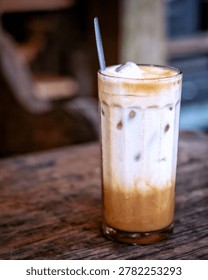 This high-quality image features a beautifully layered iced latte or iced cappuccino served in a tall, faceted clear glass. The drink showcases a sophisticated ombre effect, with rich, dark espresso at the bottom gradually blending into creamy white milk and topped with a thick, velvety layer of milk foam. Visible ice cubes nestled within the glass add to the refreshing, chilled aesthetic.
A reusable metal straw is placed inside, suggesting a modern, eco-friendly lifestyle choice. The glass sits on a rustic, textured wooden table, creating a warm and inviting cafe ambiance. The background is softly blurred with a shallow depth of field, ensuring the focus remains entirely on the beverage. This versatile photograph is perfect for summer refreshment concepts, cafe menus, food blogs, or lifestyle advertisements highlighting professional barista craft and morning coffee rituals.