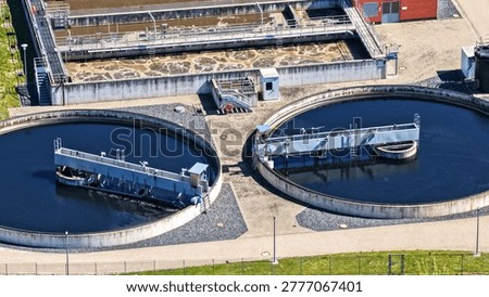 This high-resolution aerial photograph provides a detailed close-up view of a modern wastewater treatment plant in operation. The image prominently features two large circular secondary clarifiers equipped with rotating bridge scrapers and skimmers, alongside rectangular aeration and sedimentation basins with visible surface agitation and brown foam typical of the biological treatment process.
The technical infrastructure, including walkways, metal platforms, pipes, ladders, and monitoring equipment, is clearly visible. The dark blue water in the clarifiers contrasts strongly with the turbulent, foamy surface of the rectangular tanks, illustrating the different stages of the wastewater purification process.
This image is ideal for illustrating the functional and technical aspects of municipal sewage treatment. It shows key components such as final clarifiers, sludge scrapers, aeration systems, and plant operation in detail. Captured from above on a sunny day, the clear shadows emphasize the three-dimensional structure of the facility and the engineering precision involved.
Perfect for editorial use in engineering magazines, technical reports, environmental studies, educational materials, websites of water utilities, and presentations on sustainable water management, circular economy, and environmental technology. The photo effectively demonstrates how modern wastewater treatment plants clean water through mechanical and biological processes before returning it safely to the environment.