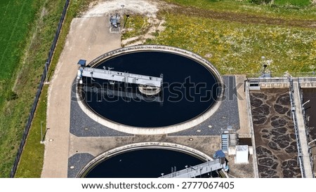 This high-resolution aerial photograph provides a detailed close-up view of a modern wastewater treatment plant in operation. The image prominently features two large circular secondary clarifiers equipped with rotating bridge scrapers and skimmers, alongside rectangular aeration and sedimentation basins with visible surface agitation and brown foam typical of the biological treatment process.
The technical infrastructure, including walkways, metal platforms, pipes, ladders, and monitoring equipment, is clearly visible. The dark blue water in the clarifiers contrasts strongly with the turbulent, foamy surface of the rectangular tanks, illustrating the different stages of the wastewater purification process.
This image is ideal for illustrating the functional and technical aspects of municipal sewage treatment. It shows key components such as final clarifiers, sludge scrapers, aeration systems, and plant operation in detail. Captured from above on a sunny day, the clear shadows emphasize the three-dimensional structure of the facility and the engineering precision involved.
Perfect for editorial use in engineering magazines, technical reports, environmental studies, educational materials, websites of water utilities, and presentations on sustainable water management, circular economy, and environmental technology. The photo effectively demonstrates how modern wastewater treatment plants clean water through mechanical and biological processes before returning it safely to the environment.