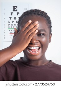 This high-resolution image features a cheerful young Black woman undergoing a vision assessment. She is playfully covering her right eye with her hand while smiling broadly, showcasing a sense of positivity and comfort during a medical checkup. In the background, a Snellen eye chart is clearly visible, though slightly out of focus, establishing a clinical yet approachable setting. The lighting is bright and even, emphasizing her clear skin and the authentic joy of her expression.