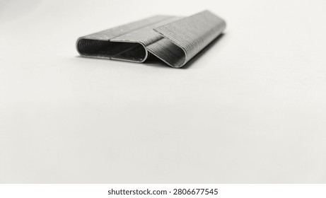 This high-resolution photograph features two strips of metal staples, precisely cut and prepared for industrial or heavy-duty use. The perspective is a three-quarter view from the top front, showing the length, the top channel, and the segmented texture of the individual staple wires fused into a strip. The two strips are positioned slightly overlapping and angled towards each other, resting on a clean, seamless white background. This arrangement creates depth and clearly showcases the shape, size, and metallic material of the product. The staples are made of galvanized steel, giving them a distinct industrial sheen and a textured, repeating pattern. The lighting is soft and even, highlighting the metallic surface and cast delicate shadows. The product is in new condition, clean and ready for use. This precise product shot is suitable for catalog listings, construction and hardware related content, or as a general tool and supply image. [1048 karakter]