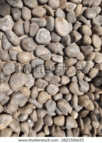 This high-resolution, top-down photograph captures a beautiful and dense arrangement of natural river stones, creating a perfect organic background. The image showcases a meticulous composition of various smooth, rounded pebbles and cobbles in a harmonious palette of grey, charcoal, earthy brown, and subtle beige tones. Each stone features a unique shape, from oval to spherical, sculpted naturally by centuries of water erosion. The gentle, natural sunlight hits the surface of the stones, highlighting their fine matte textures, subtle mineral veins, and soft contours, adding depth and a tactile dimension to the overall image.This versatile stock photo is an ideal choice for a wide variety of creative projects. It perfectly symbolizes concepts of zen, mindfulness, meditation, tranquility, and natural balance, making it excellent for wellness, spa, and healthcare branding. Additionally, it serves as a high-quality texture and pattern for architectural renderings, interior design concepts, outdoor landscaping projects, and eco-friendly or sustainable product packaging. The neutral color scheme ensures it can be used seamlessly as a website background, banner, or social media backdrop without overpowering text overlay. Whether you are looking for a rustic, rugged outdoor feel or a clean, minimalist Japanese garden aesthetic, this premium stone texture offers the perfect visual solution for graphic designers, content creators, and marketers alike.