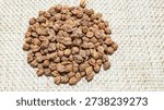 This high-resolution, top-down shot features a neatly arranged pile of raw, dry brown chickpeas, commonly known as Kala Chana or Desi Chana. The legumes are placed on a light-colored, textured jute burlap surface, which provides a rustic and organic feel to the composition. Each chickpea displays its natural wrinkled skin and earthy brown tones, emphasizing its high fiber and protein content. This image is perfect for culinary blogs, healthy eating guides, Indian or Middle Eastern recipe websites, agricultural marketing, and grocery store branding. The neutral color palette and clean arrangement make it ideal for use as a background or as a focal point for food packaging design.