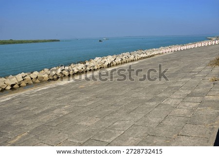 This image was captured along the eastern bank of the Bakkhali River in Cox’s Bazar, Bangladesh, directly opposite the estuary where the river meets the Bay of Bengal. The area is strongly influenced by tidal fluctuations, storm surges, and cyclonic weather systems, which cause sudden rises in water level during high tide and extreme weather events.

Large concrete blocks are installed along the riverbank as part of a city-protective embankment system designed to prevent erosion, reduce wave impact, and protect urban infrastructure from flooding. Beneath the concrete blocks, soil compaction is applied to stabilize the foundation, and geotextile fabric is used to prevent soil displacement while allowing controlled water filtration.

The concrete blocks are placed in a geometric arrangement to dissipate wave energy and minimize hydraulic pressure during tidal surges and storm conditions. This type of engineered shoreline protection is commonly used along tidal rivers and coastal estuarine zones to safeguard cities, roads, and public spaces from erosion and flood damage. The scene represents coastal and riverbank protection infrastructure under clear daylight conditions.