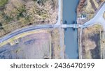This image captures an aerial perspective of a canal intersecting with a road. The photograph depicts a controlled water passage with a lock system, allowing boats to navigate between different water