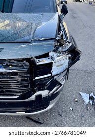 This image captures the aftermath of a collision between two vehicles, showing the damaged front end of an SUV. The hood is crumpled and dislocated, with the front grill and headlight assembly shattered and partially detached from the body of the vehicle. Various broken plastic and metal pieces are scattered on the ground around the SUV, reflecting the intensity of the impact. The cracked headlight is barely intact, and the windshield shows signs of being affected by the crash. The vehicle is parked on a road, with other cars visible in the background, indicating this incident took place in an urban environment. The scene portrays the severity of the damage, highlighting the need for immediate repairs. The image evokes a sense of shock and the dangerous consequences of road accidents. The lighting reflects the clarity of the moment, focusing on the crushed parts and highlighting the extent of the damage. The SUV's black and silver paint is still visible under the debris, giving a stark contrast to the broken components.