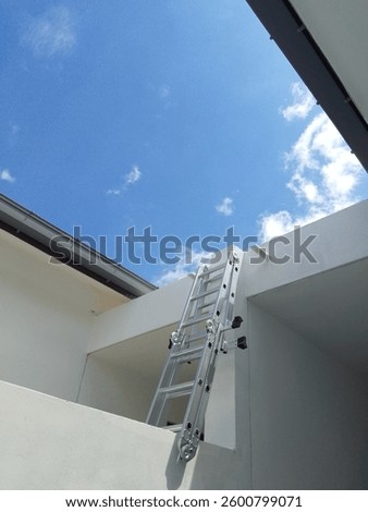This image captures an aluminum multipurpose ladder leaning against a white modern house under a bright blue sky with scattered clouds. The architectural design features clean lines and minimalistic.