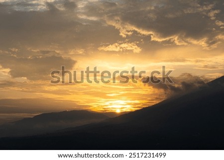 This image captures a beautiful sunset over a mountain range. The sky is a vibrant orange and yellow, with cloudy sky. The sun is setting behind the mountains, casting long shadows over the landscape.