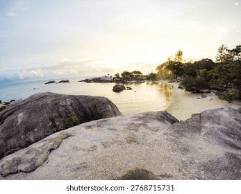 This image captures a breathtaking beach scene at sunrise, with large rocks and lush greenery in the foreground, overlooking a calm ocean. The serene atmosphere and tranquil waters create a sense of seclusion and paradise, perfect for a relaxing getaway or adventure. The rocky shoreline and surrounding ecosystem add to the natural beauty of the scene, highlighting the importance of conservation and sustainability. The location of this stunning scene appears to be a tropical island, possibly in the Seychelles or similar archipelago.