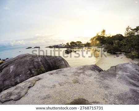 This image captures a breathtaking beach scene at sunrise, with large rocks and lush greenery in the foreground, overlooking a calm ocean. The serene atmosphere and tranquil waters create a sense of seclusion and paradise, perfect for a relaxing getaway or adventure. The rocky shoreline and surrounding ecosystem add to the natural beauty of the scene, highlighting the importance of conservation and sustainability. The location of this stunning scene appears to be a tropical island, possibly in the Seychelles or similar archipelago.