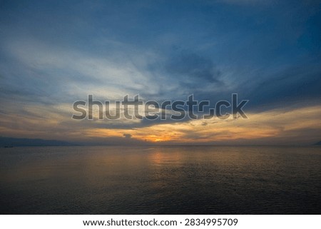 This image captures a breathtaking sunset over a calm sea, where the horizon glows with warm shades of orange and gold fading into soft blues and grays. The sky is filled with delicate, wispy clouds that reflect the fading sunlight, creating a peaceful and atmospheric composition. The gentle ripples on the water mirror the colors of the sky, adding depth and tranquility to the scene. The distant silhouettes of landmasses enhance the sense of scale and serenity. This photo beautifully conveys the quiet elegance of nature at dusk, making it ideal for use in travel, landscape, and nature-themed projects. It evokes feelings of calm, reflection, and timeless beauty — a perfect representation of the harmony between sea and sky.