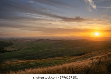 This image captures the breathtaking view of a sunset over rolling hills and fields in Tuscany. The warm hues of the setting sun cast long shadows across the landscape, creating a serene atmosphere. A lone tree stands as a silhouette against the vibrant sky. Tuscany, Asciano, Italy