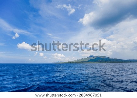 This image captures a breathtaking view of the open sea under a bright blue sky scattered with soft white clouds. The deep blue ocean stretches toward a distant coastline lined with green hills and mountains, creating a perfect harmony between land, sea, and sky. The gentle waves reflect the sunlight, adding texture and movement to the serene scene. The composition evokes feelings of freedom, tranquility, and natural beauty — ideal for themes related to travel, adventure, and tropical landscapes. The vivid colors and expansive horizon make this photo perfect for use in tourism, environmental, or nature-inspired projects, symbolizing peace and the vastness of the ocean.