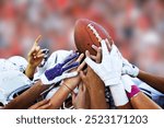 This image captures a close-up moment of a group of American football players lifting a football together, symbolizing unity and team spirit. The players, wearing helmets and gloves.