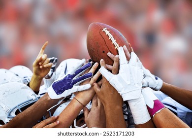 This image captures a close-up moment of a group of American football players lifting a football together, symbolizing unity and team spirit. The players, wearing helmets and gloves. - Powered by Shutterstock