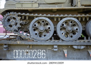 This image captures a close-up perspective of a heavy military vehicle's rugged drive mechanism, highlighting its metallic tracks and wheels. The scene displays the lower section of a tracked transport unit, focusing on the mechanical complexity of its propulsion system. On the left, a large, toothed sprocket gear engages with the thick, articulated links of the steel treads, which appear weathered from past operational use. Adjacent to this drive component, two primary road wheels feature circular cutouts and are finished in a dark, matte grey paint that shows signs of minor surface oxidation and wear. The machinery rests upon a sturdy, flat bed structure, painted in a similar olive-drab tone, which bears a prominent, bold identifier printed in large white digits reading 00000286. A small, triangular orange marker is attached near the bottom edge of this platform, providing a sharp contrast to the surrounding muted color palette. The composition emphasizes the industrial aesthetic and functional design of the heavy-duty assembly, showcasing textures ranging from rough, rusted metal to smooth, cast surfaces. Every component contributes to the impression of immense weight and durability, with bolts, hinges, and structural support brackets adding layers of intricate detail to the engineering display. Faint traces of green paint remain on the chassis above the tracks, hinting at the original camouflage or protective coating of the unit, while the shadows beneath the wheels and between the gear teeth suggest a deep, complex geometry beneath the exterior plating. The entire arrangement serves as an evocative study of military hardware, frozen in a static moment of repose while mounted on its transport platform, waiting for potential deployment or exhibition.