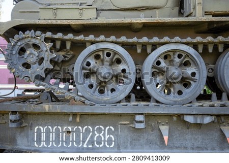 This image captures a close-up perspective of a heavy military vehicle's rugged drive mechanism, highlighting its metallic tracks and wheels. The scene displays the lower section of a tracked transport unit, focusing on the mechanical complexity of its propulsion system. On the left, a large, toothed sprocket gear engages with the thick, articulated links of the steel treads, which appear weathered from past operational use. Adjacent to this drive component, two primary road wheels feature circular cutouts and are finished in a dark, matte grey paint that shows signs of minor surface oxidation and wear. The machinery rests upon a sturdy, flat bed structure, painted in a similar olive-drab tone, which bears a prominent, bold identifier printed in large white digits reading 00000286. A small, triangular orange marker is attached near the bottom edge of this platform, providing a sharp contrast to the surrounding muted color palette. The composition emphasizes the industrial aesthetic and functional design of the heavy-duty assembly, showcasing textures ranging from rough, rusted metal to smooth, cast surfaces. Every component contributes to the impression of immense weight and durability, with bolts, hinges, and structural support brackets adding layers of intricate detail to the engineering display. Faint traces of green paint remain on the chassis above the tracks, hinting at the original camouflage or protective coating of the unit, while the shadows beneath the wheels and between the gear teeth suggest a deep, complex geometry beneath the exterior plating. The entire arrangement serves as an evocative study of military hardware, frozen in a static moment of repose while mounted on its transport platform, waiting for potential deployment or exhibition.
