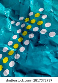 This image captures a closeup view of a crumpled piece of teal paper. The paper features an arrangement of white and yellow oval shapes, possibly representing a decorative pattern or a stylized element. The texture of the paper, with its folds and creases, adds a tactile dimension to the visual. The lighting creates highlights and shadows, emphasizing the threedimensional quality of the surface.