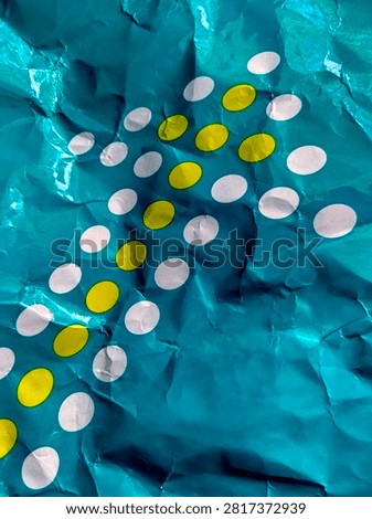 This image captures a closeup view of a crumpled piece of teal paper. The paper features an arrangement of white and yellow oval shapes, possibly representing a decorative pattern or a stylized element. The texture of the paper, with its folds and creases, adds a tactile dimension to the visual. The lighting creates highlights and shadows, emphasizing the threedimensional quality of the surface.
