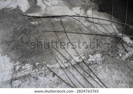 This image captures a close-up view of an aged, cracked pavement surface, likely asphalt or concrete. The surface shows extensive wear, with numerous intersecting fissures and uneven patches. Overhead, shadows from metal beams or structural supports are cast onto the pavement, creating stark contrasts and adding depth to the scene. The lighting suggests it might be taken during the early morning or late afternoon, emphasizing the texture and deterioration of the material. The overall scene evokes a sense of urban decay and the natural aging process of infrastructure.