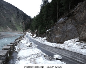 This image captures a dramatic mountain road, still partially covered with significant snowdrifts from winter, winding alongside a flowing river in the high altitudes of northern Pakistan