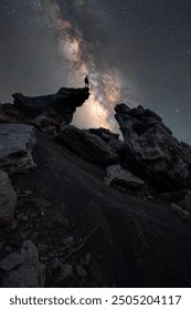 This image captures a dramatic night sky scene featuring a person standing on a rocky outcrop, silhouetted against the Milky Way. The stars and the vibrant colors of the galaxy form a breathtaking bac