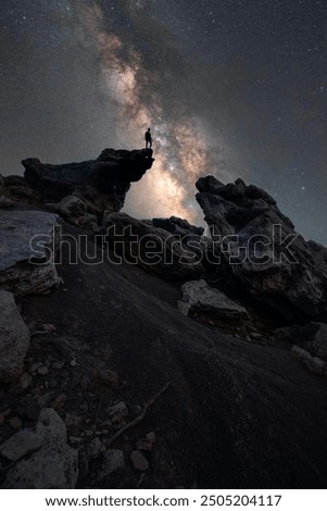 This image captures a dramatic night sky scene featuring a person standing on a rocky outcrop, silhouetted against the Milky Way. The stars and the vibrant colors of the galaxy form a breathtaking bac