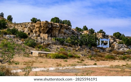 This image captures Eastern Pageant Canyon in the northwestern part of Cimarron County, Oklahoma. The area is known for annual theatrical performances reenacting the Easter story. In the background are characteristic sandstone rock formations typical of the region. A cholla grows in the left foreground, while the landscape consists of dry grass and rocky hills.