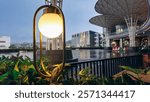 This image captures a glowing outdoor lamp surrounded by lush greenery at Solaria in Lampung City Mall, with modern architecture and a serene water feature in the background.

