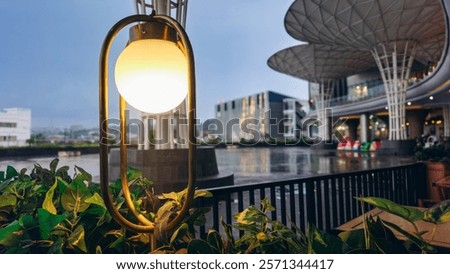 This image captures a glowing outdoor lamp surrounded by lush greenery at Solaria in Lampung City Mall, with modern architecture and a serene water feature in the background.

