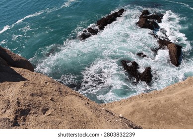 This image captures a high-angle, downward perspective looking over the edge of a rugged sandstone cliff toward a rocky coastline. Below, turbulent turquoise ocean water swirls and crashes violently around a cluster of dark, jagged reef rocks.