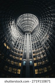 This image captures the interior of a striking modern building characterized by its expansive, circular glass ceiling. The ceiling, composed of a grid of glass panels, allows natural light to flood the space, creating a bright and airy atmosphere. The central support column, extending from the base to the top, adds a sense of verticality and structural integrity. The surrounding walls feature numerous windows, offering glimpses of the exterior while allowing ample daylight to permeate the interior. The overall design exudes a sense of openness and sophistication, making it a remarkable example of contemporary architecture.